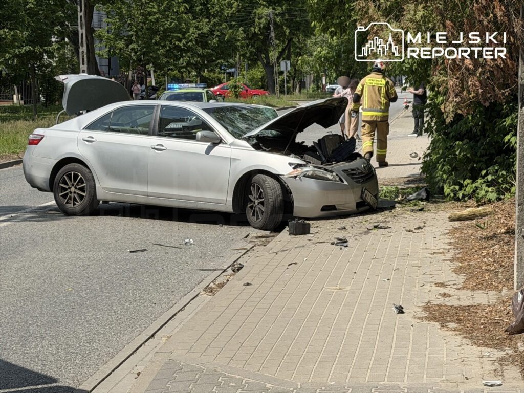 Uszkodzony samochód osobowy z otwartą maską stoi na ulicy, obok pracują strażacy oraz zebrani przechodnie.