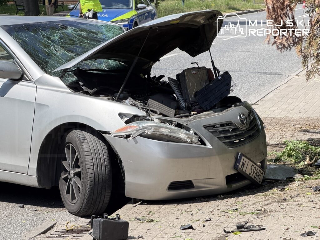 Uszkodzony samochód osobowy z otwartą maską stoi na chodniku obok drogi, w tle widać radiowóz policyjny.