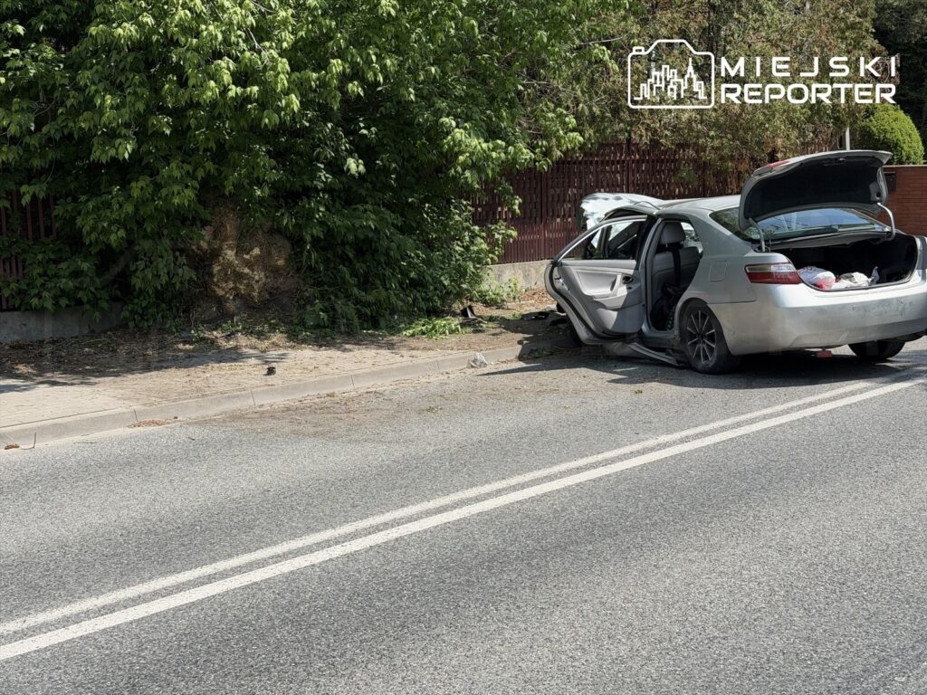 Uszkodzony samochód zaparkowany przy drodze, z otwartymi drzwiami i bagażnikiem, otoczony zielenią.