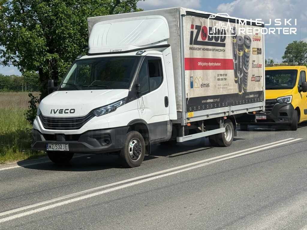 Biały samochód dostawczy marki Iveco zatrzymany na drodze, obok żółty pojazd, w tle zielone drzewa i błękitne niebo.