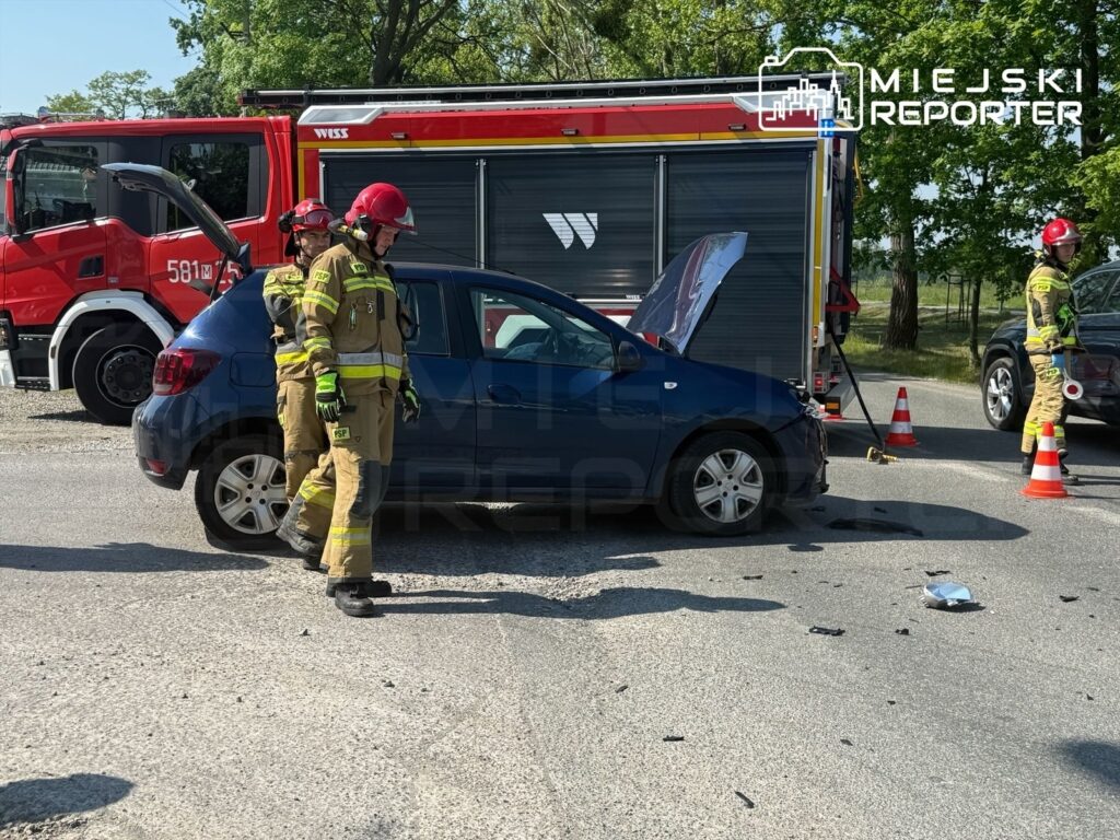 Dwaj strażacy w mundurach pracują przy niebieskim samochodzie osobowym z otwartą klapą bagażnika na drodze.