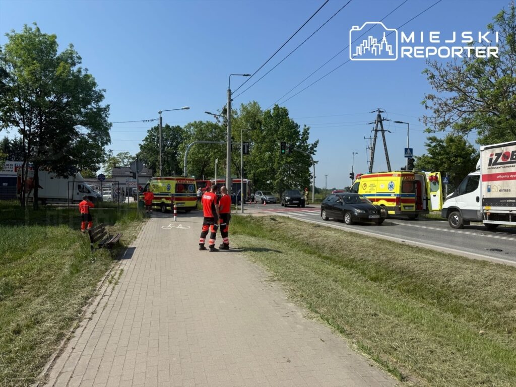 Zespół ratowników medycznych w czerwonych strojach pracuje przy drodze, obok ambulansu na sygnale.
