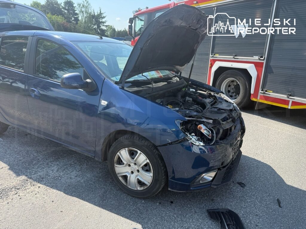 Nieprzytomny niebieski samochód z otwartą maską stoi na drodze, obok widać wóz strażacki.