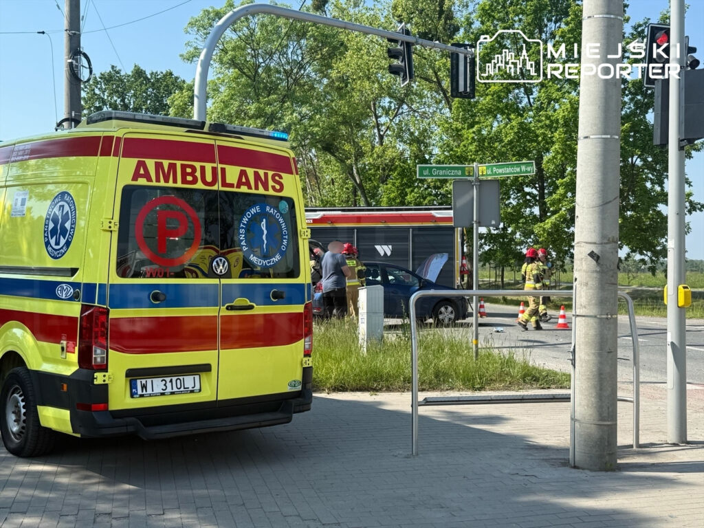 Ambulans z napisem "AMBULANS" stoi na poboczu, a obok strażacy w hełmach pracują przy wypadku drogowym.