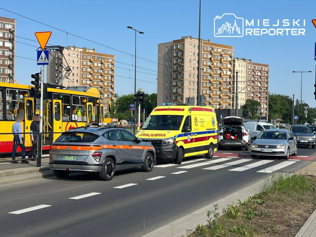 Ambulans z włączonymi sygnałami świetlnymi zatrzymany na skrzyżowaniu, obok tramwaj i pojazdy osobowe w ruchu.