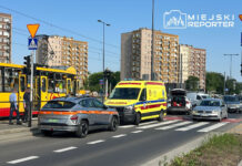 Ambulans z włączonymi sygnałami świetlnymi zatrzymany na skrzyżowaniu, obok tramwaj i pojazdy osobowe w ruchu.