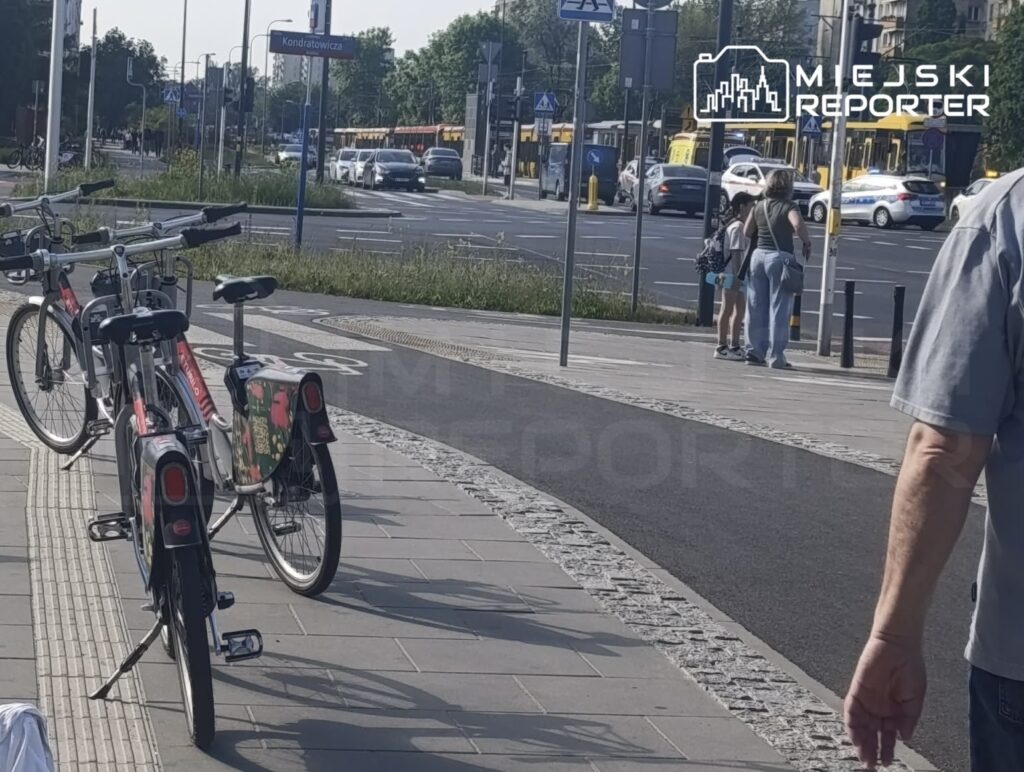 Rower miejski zaparkowany na chodniku, obok grupy ludzi czekających na przystanku w miejskim otoczeniu.