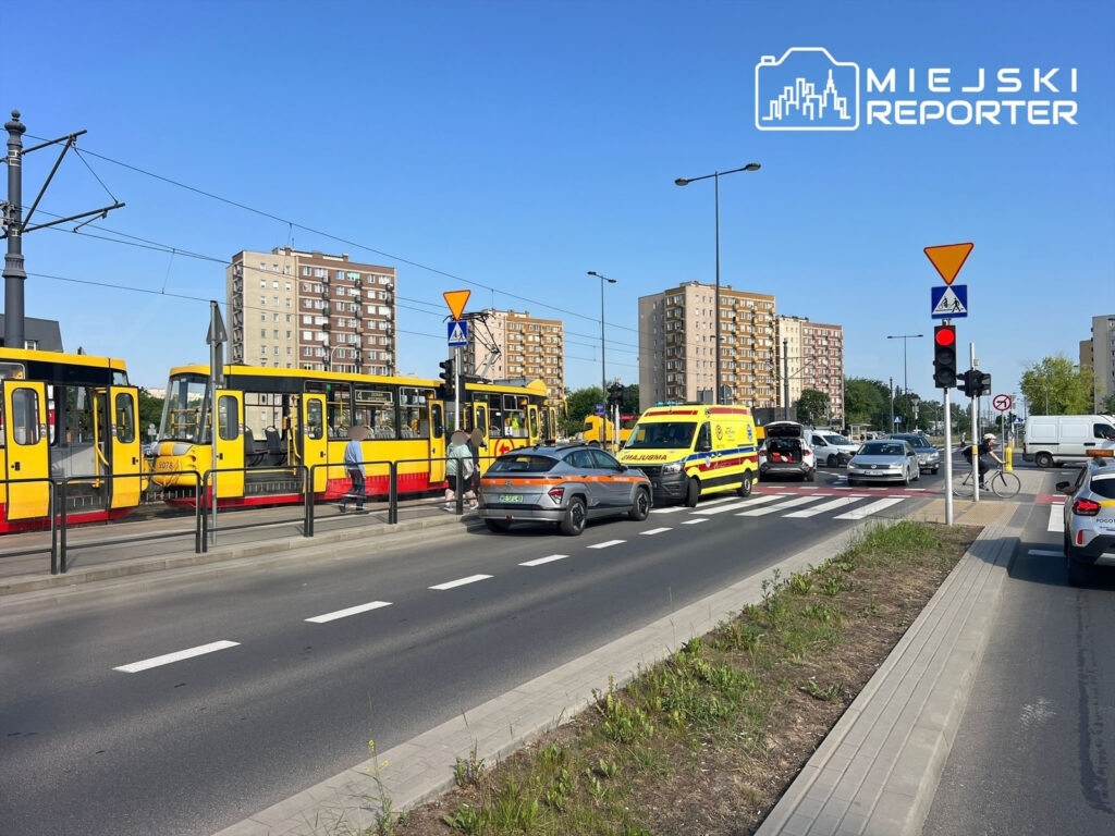 Tramwaje w żółtych barwach zatrzymane na przystanku, obok ruchliwa ulica z pojazdami i ambulans. W tle bloki mieszkalne.
