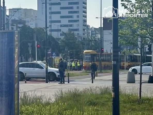Grupa osób w kamizelkach odblaskowych prowadzi prace przy torach tramwajowych w miejskim otoczeniu.