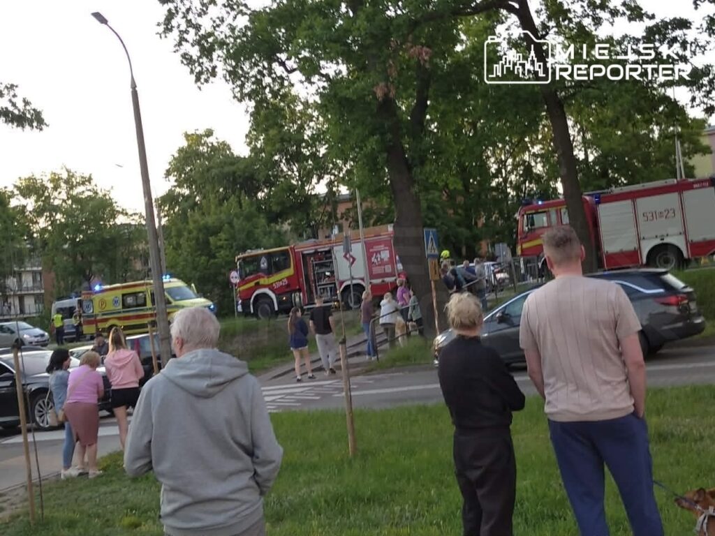 Grupa osób obserwująca działania służb ratunkowych przy drodze, obok wozów strażackich i ambulansu.