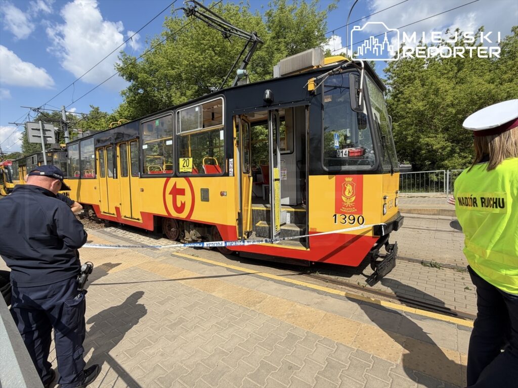 Tramwaj w kolorze żółto-czerwonym zatrzymany na przystanku, z policjantem robiącym zdjęcia i pracownikiem nadzoru ruchu w kamizelce.