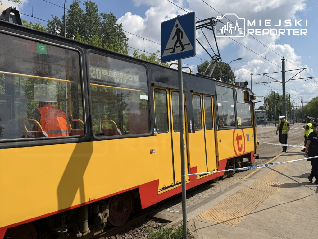 Tramwaj linii 20 zatrzymany na przystanku, obok policjanci zabezpieczający miejsce zdarzenia, w tle zielone drzewa.