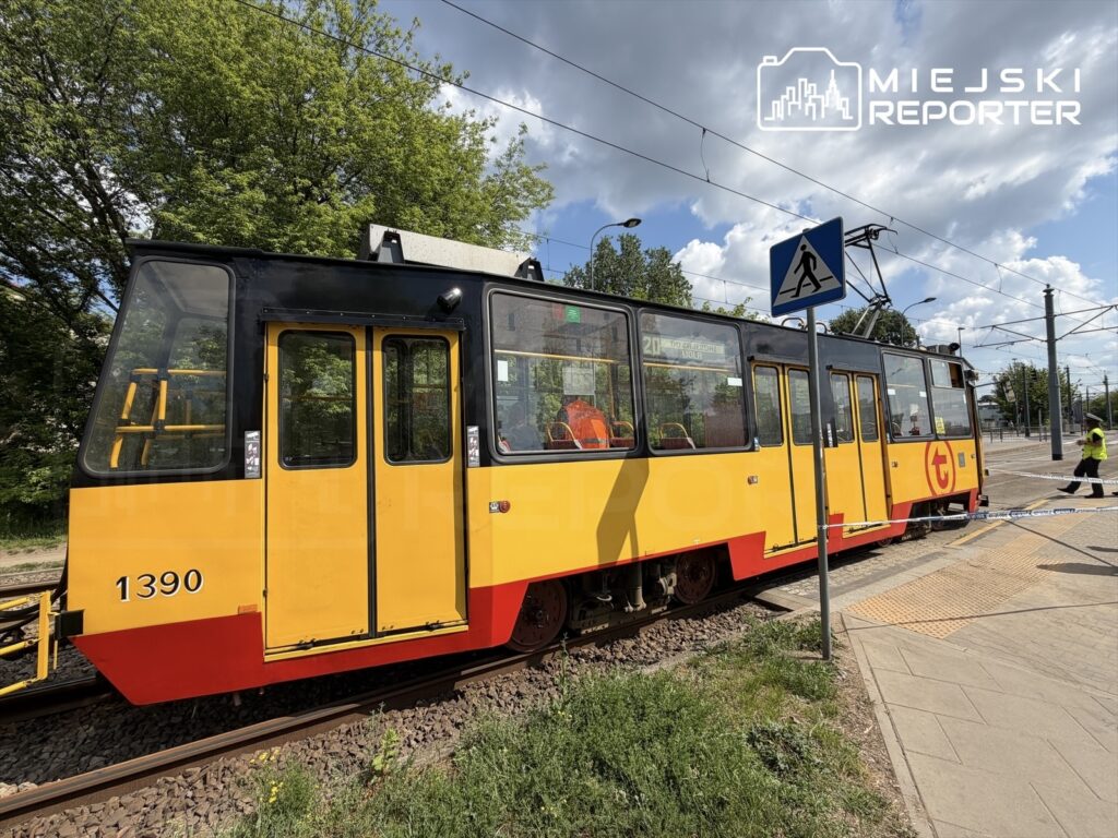 Tramwaj w żółto-czerwonym malowaniu zatrzymany na przystanku, w tle zielone drzewa i niebo z chmurami.