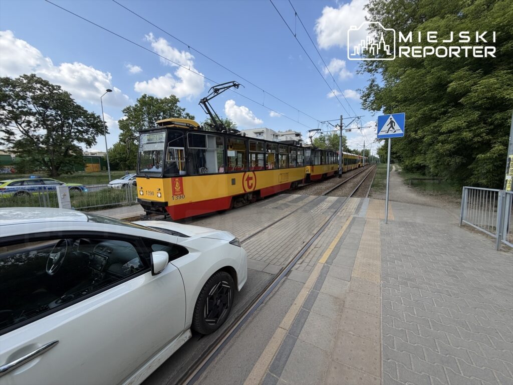 Tramwaj w żółto-czerwonym malowaniu zatrzymany na torach, obok biała osobówka na parkingu, w tle zieleń drzew.