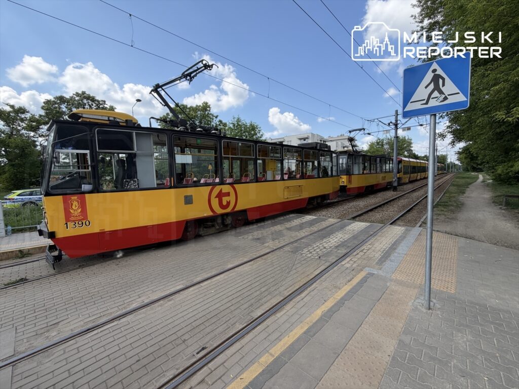 Tramwaj w żółto-czerwonym malowaniu zatrzymany na przystanku, obok znaku drogowym dla pieszych w zielonym otoczeniu.