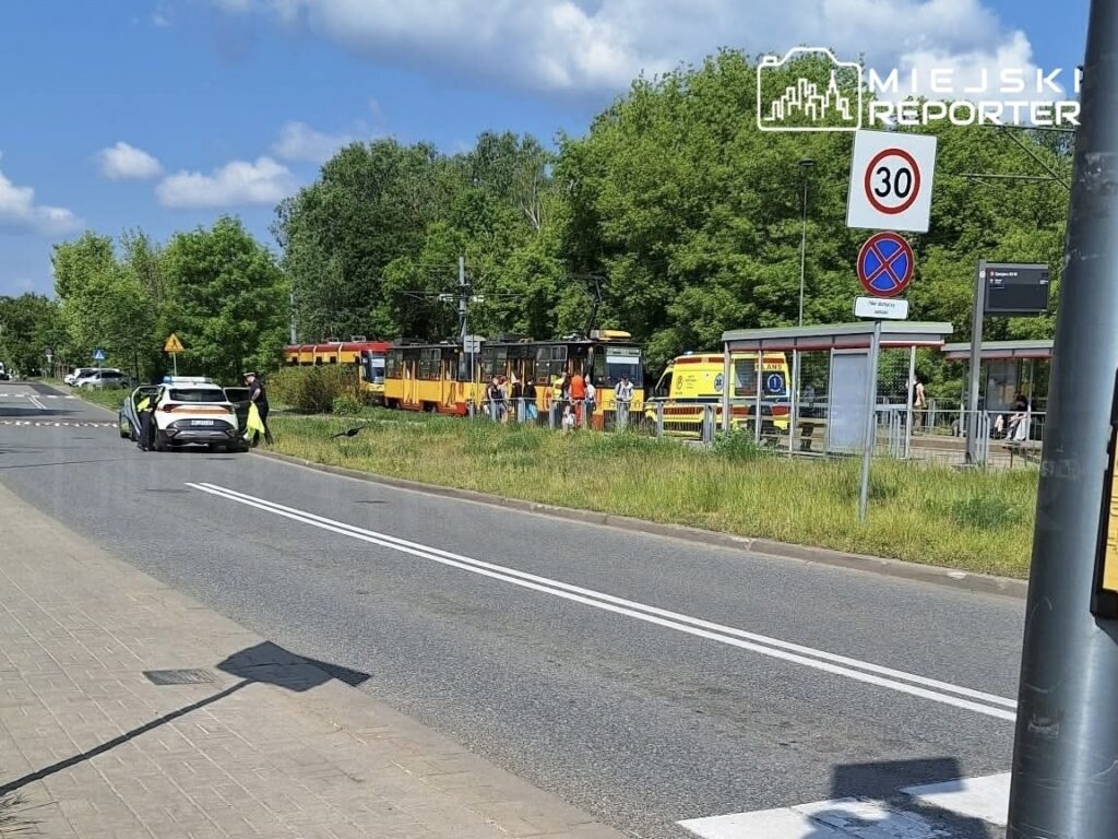 Na drodze stoi radiowóz policji, a obok grupka ludzi czeka przy przystanku tramwajowym. W tle widoczne tramwaje.