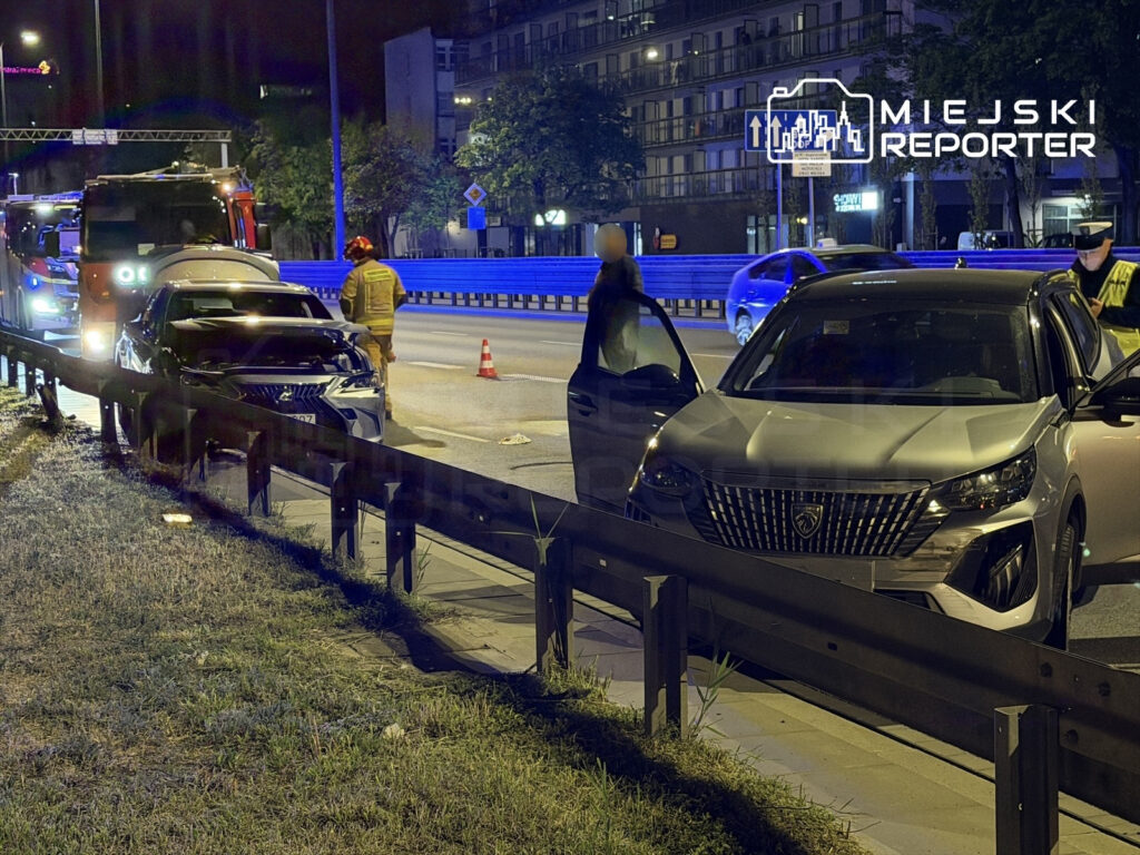 W nocy na drodze zatrzymane są dwa uszkodzone samochody, obok nich pracują strażacy i policjanci.