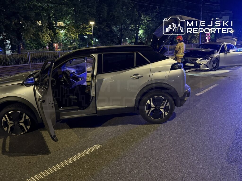 Dwa uszkodzone samochody osobowe na drodze, jeden z otwartą maską, a obok strażak w kasku. Nocna sceneria.