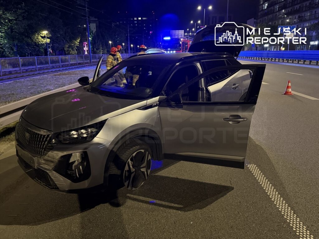 Czarny SUV z otwartymi drzwiami stoi na drodze, obok pracują strażacy w hełmach, a w tle świecą niebieskie światła.