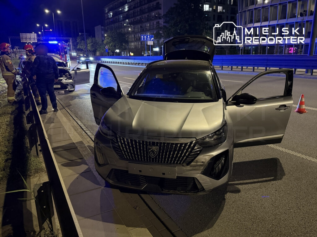 W nocy, przy drodze, strażacy udzielają pomocy osobom przy uszkodzonym samochodzie z otwartymi drzwiami.