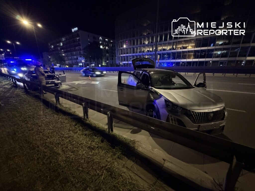 Policjanci prowadzący kontrolę drogową przy zaparkowanym samochodzie osobowym na ruchliwej ulicy w nocy.