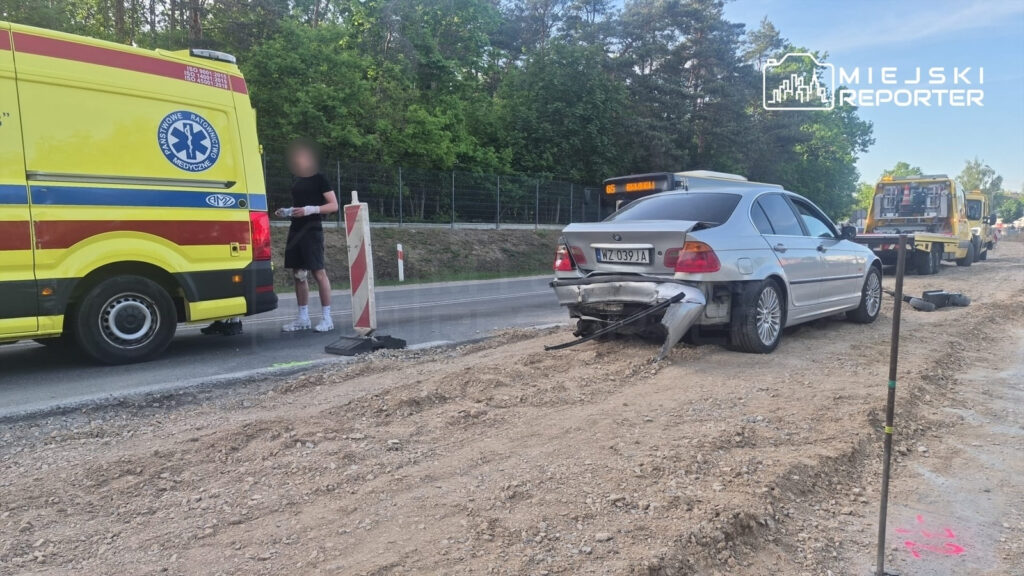 Ambulans stoi obok uszkodzonego samochodu osobowego na drodze, gdzie trwają prace budowlane. Mężczyzna w krótkich spodniach trzyma dokumenty.