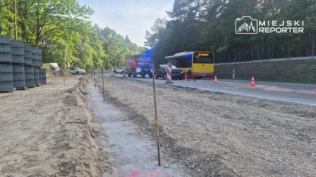 Na drodze widać nieoznakowany samochód osobowy oraz autobus, obok nich stoi wóz strażacki z włączonymi sygnałami. W tle widoczne są stosy materiałów budowlanych oraz oznaczenia robót drogowych.