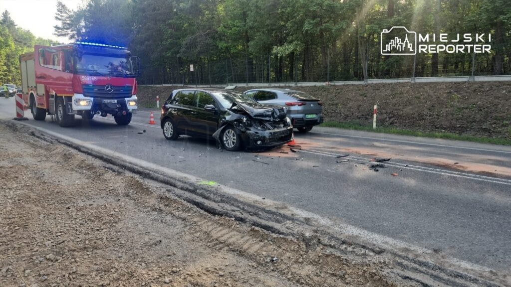 Czarny samochód osobowy z uszkodzoną przednią częścią stoi na drodze, obok niego wóz strażacki z włączonymi sygnałami.
