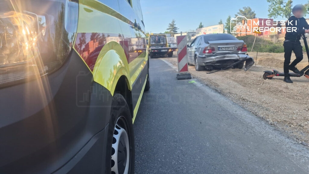 Ambulans z włączonymi światłami zatrzymany obok uszkodzonego samochodu osobowego, obok stoi mężczyzna na hulajnodze.
