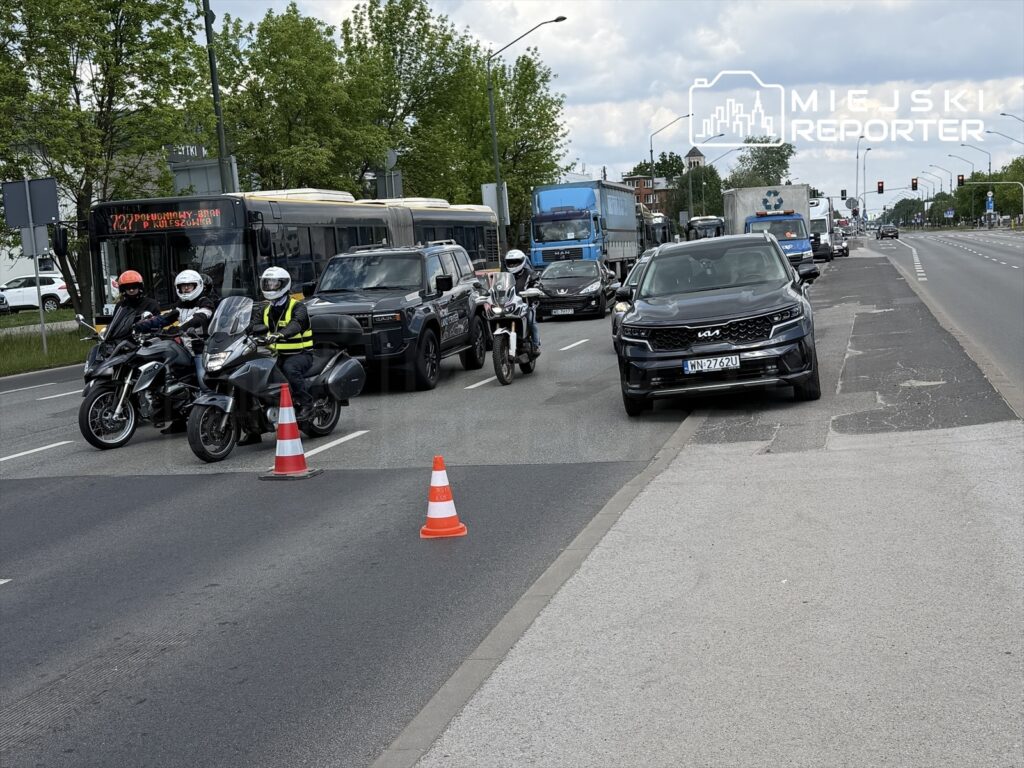 Grupa motocyklistów w kaskach zatrzymana na jezdni obok ruchliwej ulicy, z pojazdem osobowym zaparkowanym na chodniku.