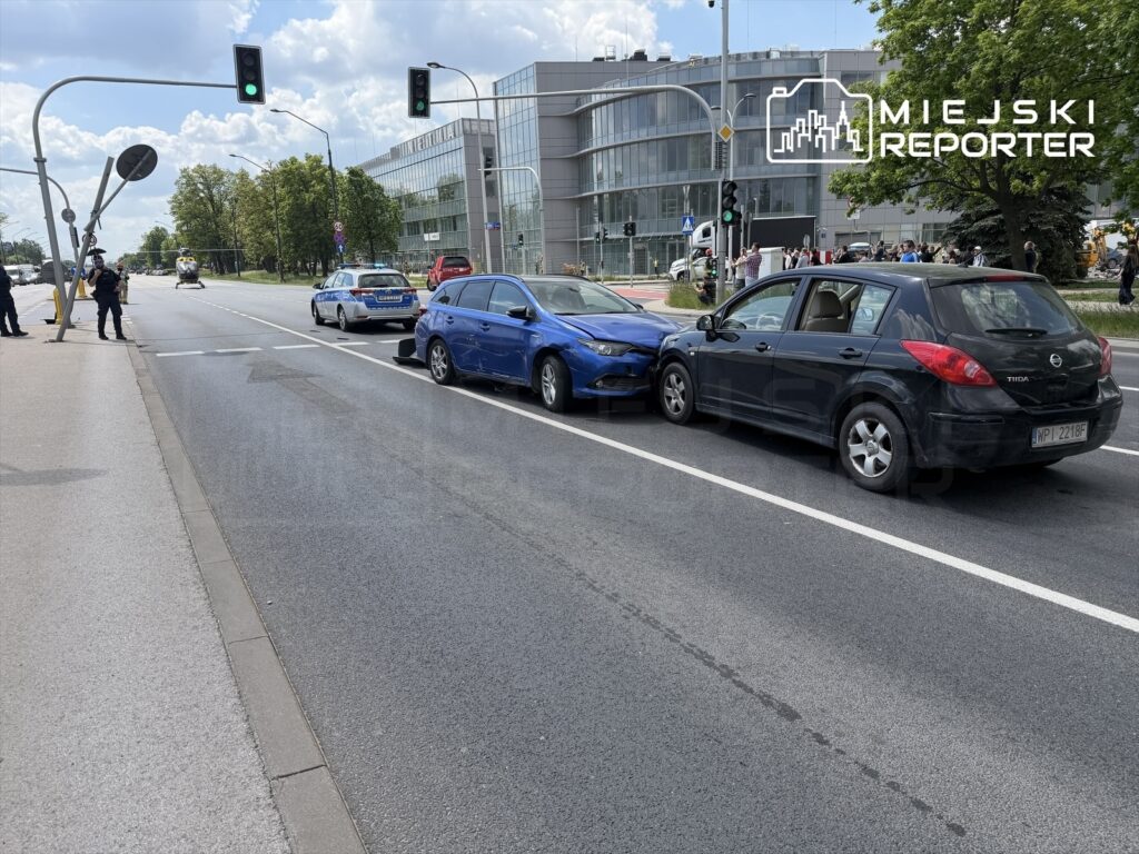 Dwa uszkodzone samochody osobowe na jezdni, obok policyjny radiowóz oraz grupa ludzi obserwujących zdarzenie.