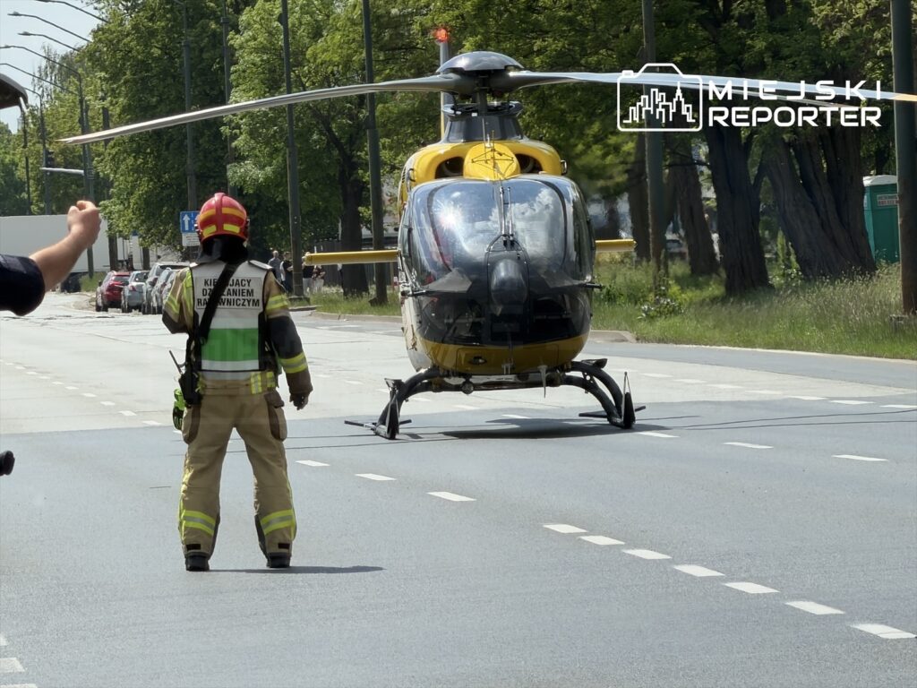 Strażak w hełmie kieruje ruchem drogowym na ulicy, obok lądującego śmigłowca ratunkowego.