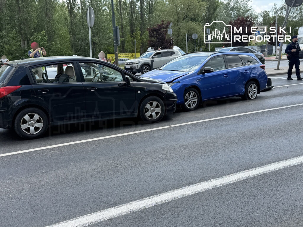 Czarne i niebieskie samochody osobowe zderzone na jezdni, obok stojący policjant oraz świadkowie zdarzenia.