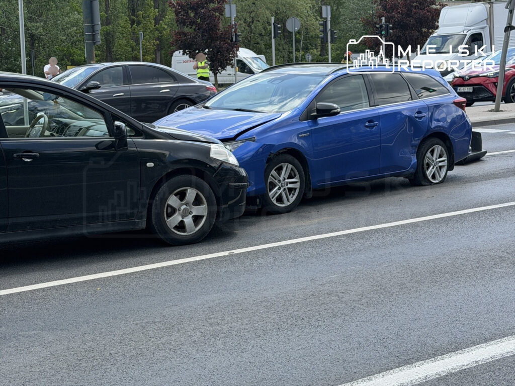 Czarny samochód osobowy zderzony z niebieskim pojazdem na miejskiej ulicy po deszczu.