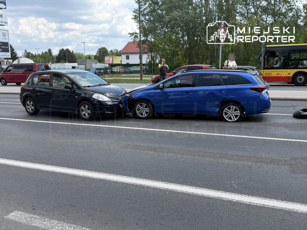 Dwa uszkodzone samochody osobowe stoją na jezdni po kolizji, obok przechodnie i ruch uliczny w tle.
