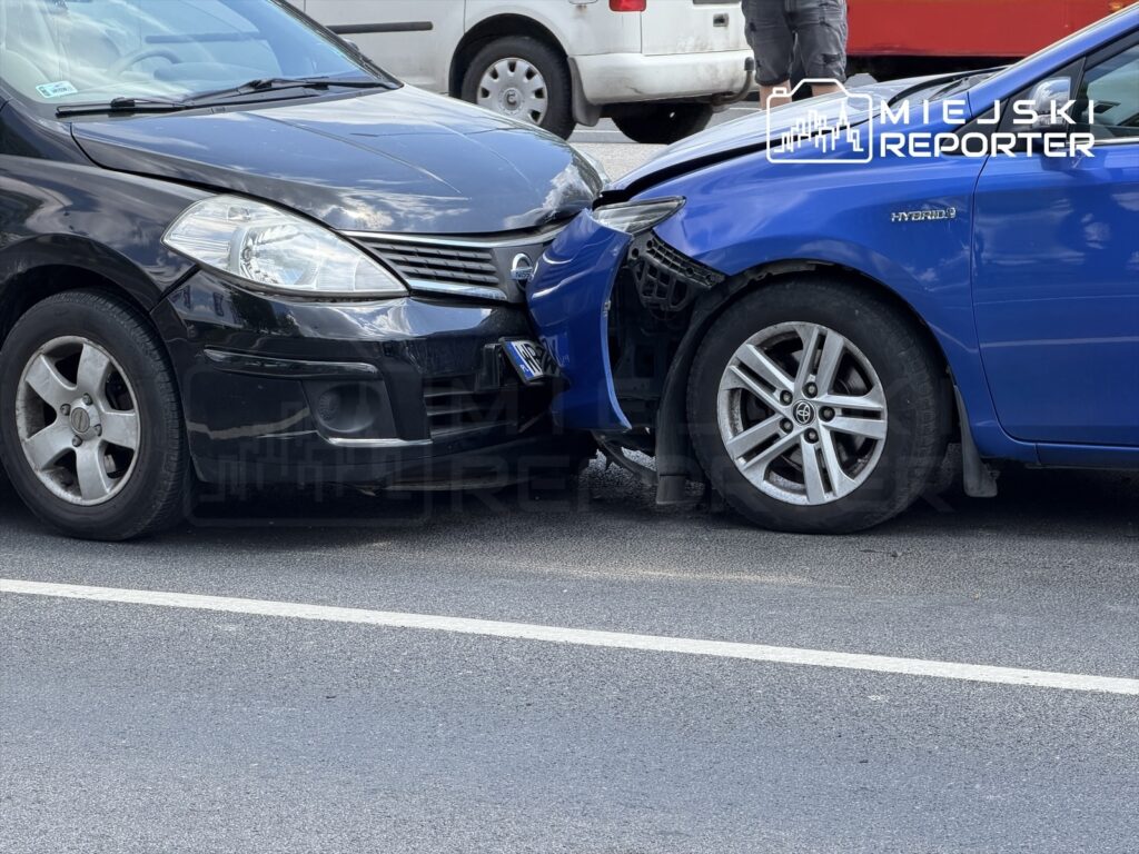 Dwa uszkodzone samochody osobowe zderzone na drodze, jeden czarny, drugi niebieski, z widocznymi uszkodzeniami karoserii.