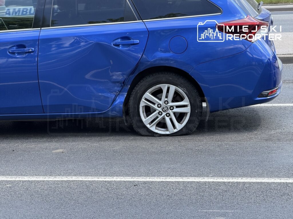Niebieski samochód osobowy z uszkodzoną karoserią i lewym tylnym kołem opadającym na asfalcie.