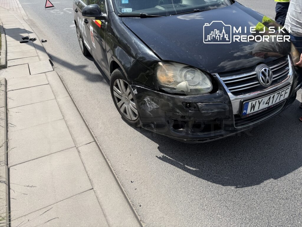 Czarny samochód osobowy marki Volkswagen z uszkodzoną przednią częścią stoi na ulicy, obok leży trójkąt ostrzegawczy.
