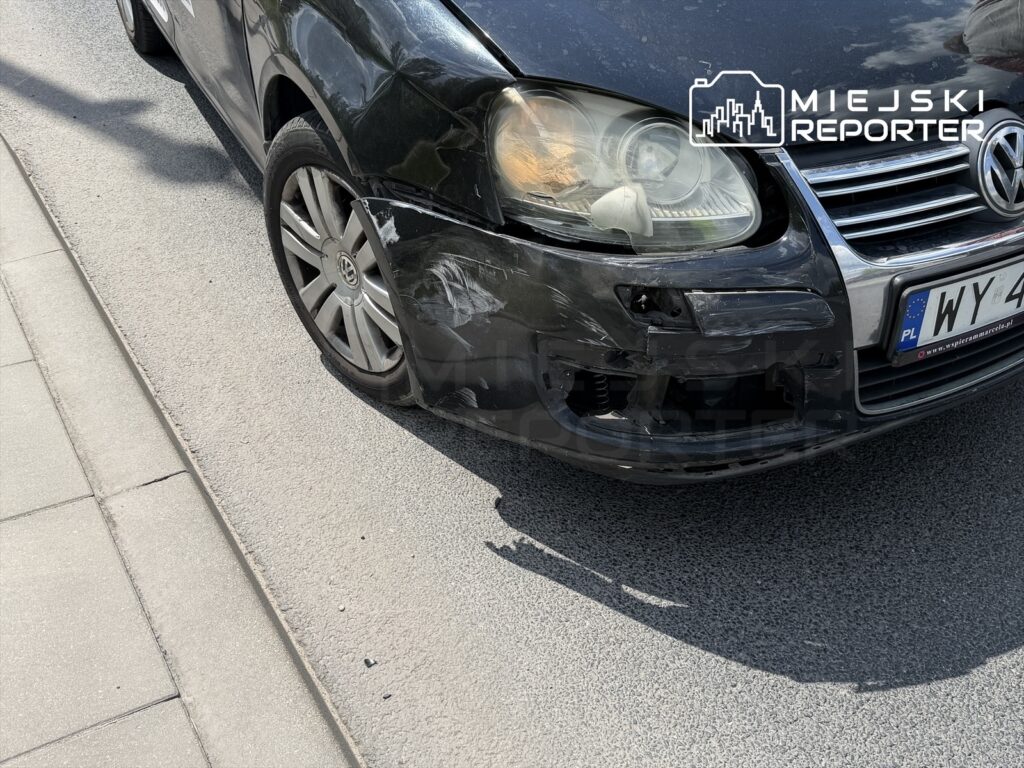 Uszkodzony czarny samochód osobowy zaparkowany na chodniku, z widocznymi zarysowaniami na przednim zderzaku.