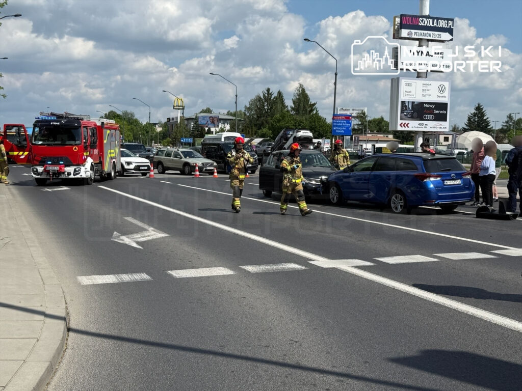 Strażacy w mundurach pracują przy zderzeniu dwóch samochodów na ruchliwej ulicy, zabezpieczając miejsce zdarzenia.