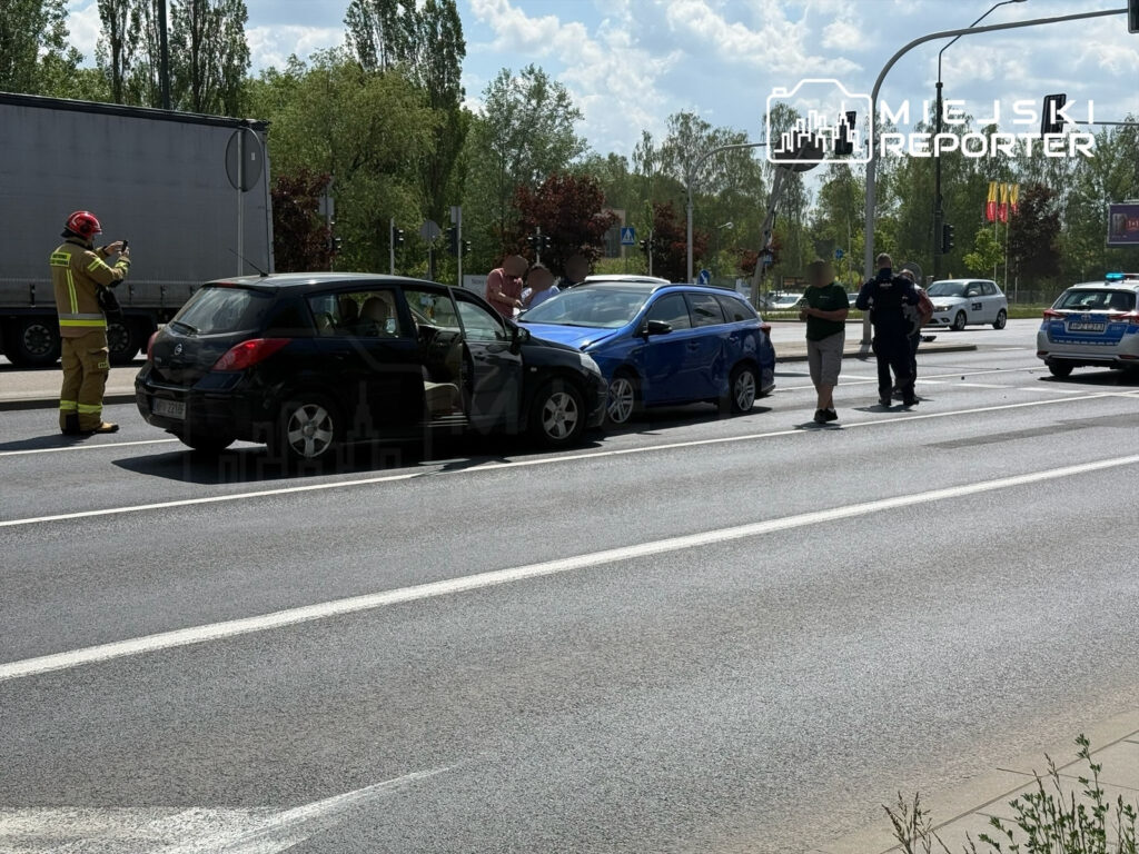 Na drodze doszło do kolizji dwóch samochodów; strażak wykonuje zdjęcia, a uczestnicy zdarzenia rozmawiają.
