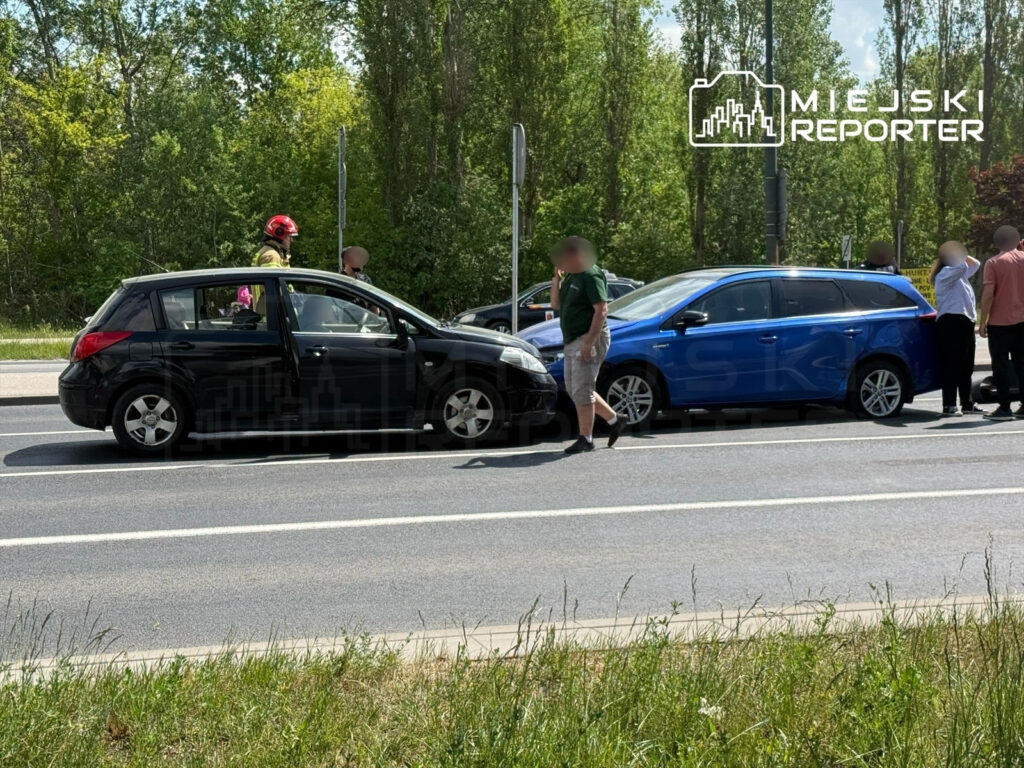 Czarny samochód osobowy stoi na drodze, obok niego grupa ludzi rozmawia z ratownikami w kaskach.