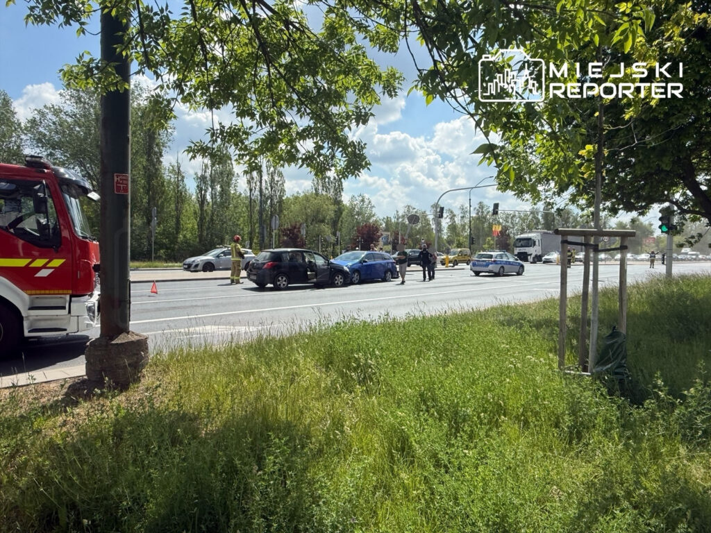 Na drodze stoją dwa uszkodzone samochody, obok nich strażak w mundurze bada sytuację, a w tle widać ruch uliczny.