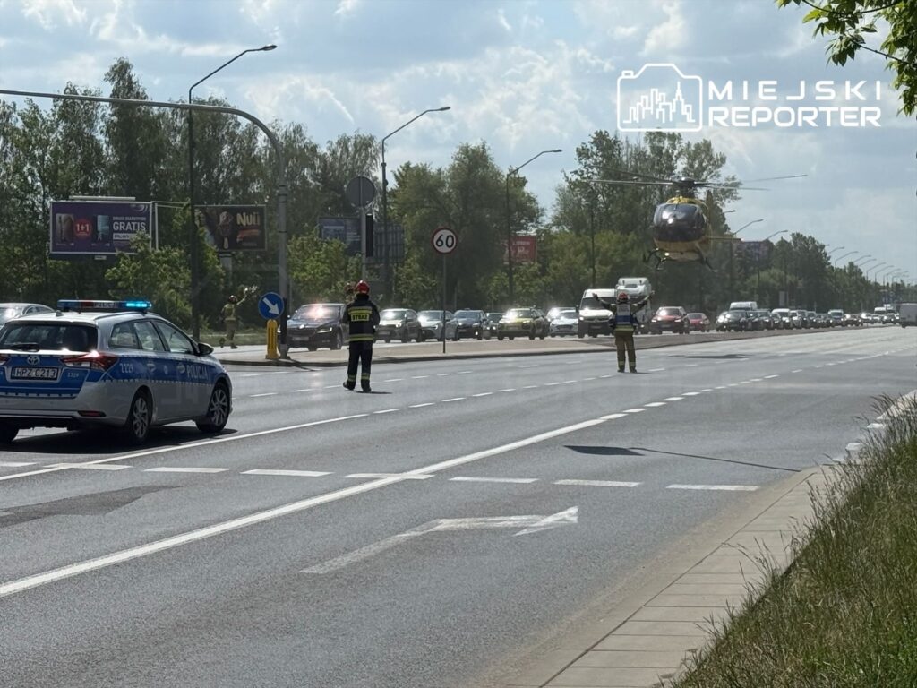 Helikopter ratunkowy unosi się nad jezdnią, obok policjanta i strażaka kierujących ruchem w mieście.