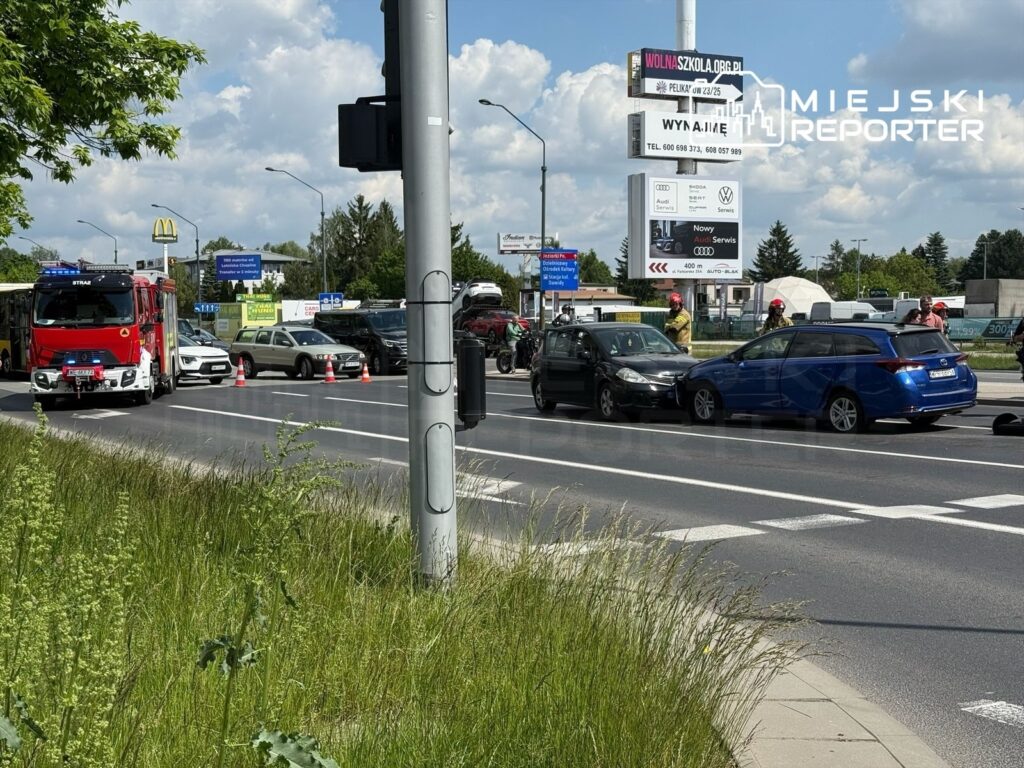 Wielu ratowników pracuje przy kolizji dwóch samochodów na ruchliwej ulicy, obok wozu strażackiego.