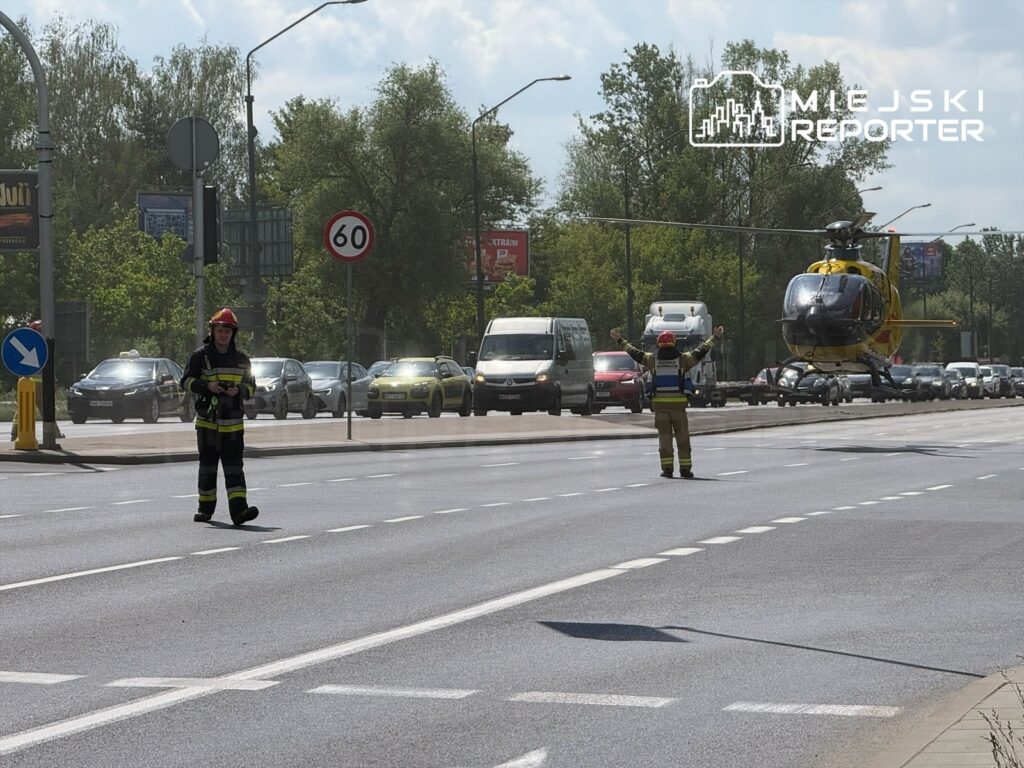 Strażak w hełmie kieruje ruchem na drodze, podczas gdy śmigłowiec unosi się nad zatorami samochodowymi.