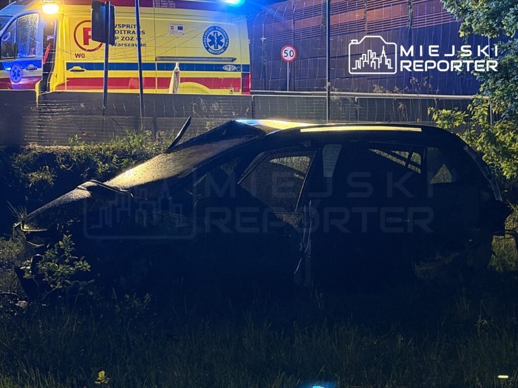 Uszkodzony samochód osobowy stoi na poboczu drogi, obok widać ambulans z włączonymi sygnałami.
