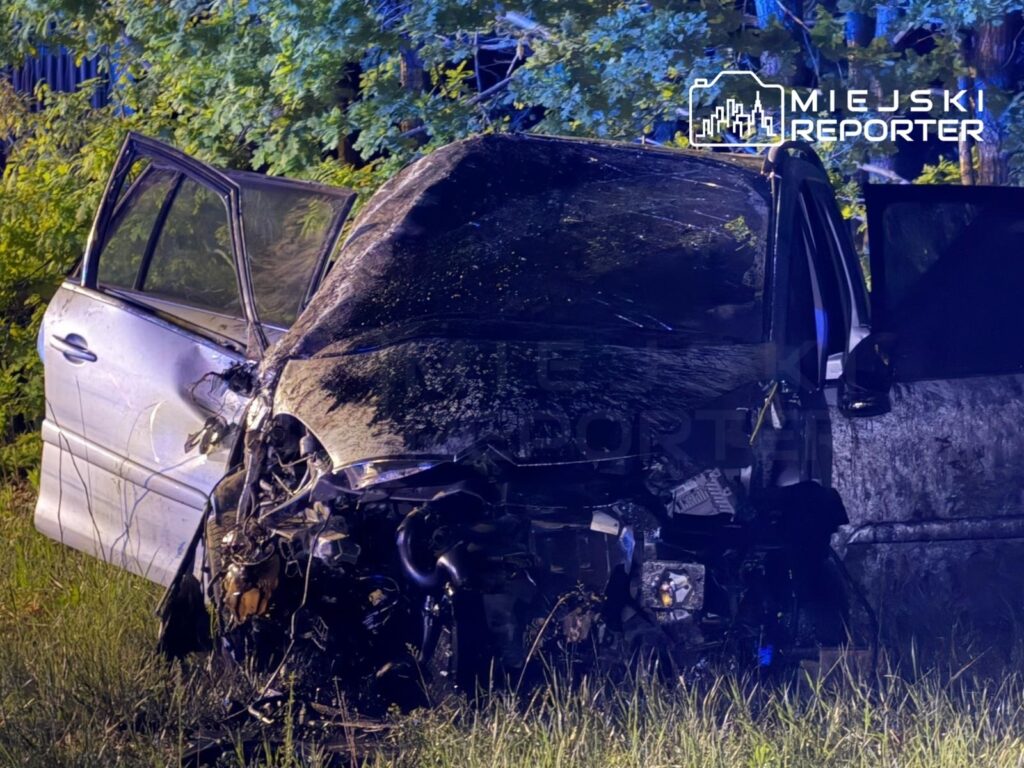 Uszkodzony samochód osobowy leży na poboczu drogi, otoczony przez zieleń, z otwartymi drzwiami po stronie kierowcy.