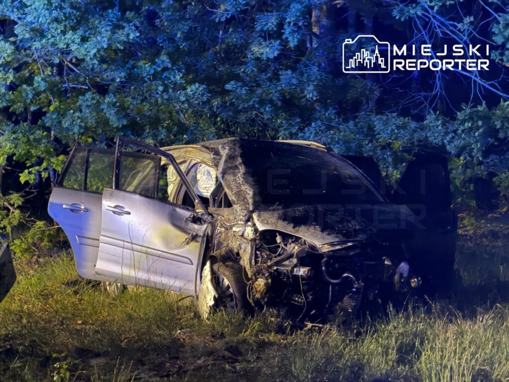Uszkodzony samochód osobowy z otwartymi drzwiami stoi na poboczu drogi, otoczony drzewami w nocy.