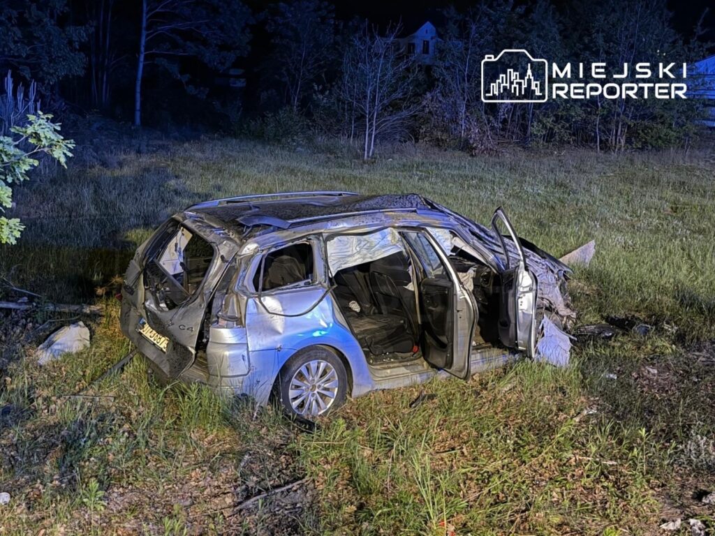Zniszczony samochód osobowy leży na poboczu drogi, otoczony trawą i drzewami, z otwartymi drzwiami.